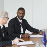 Dois homens de terno conversam à mesa durante uma reunião.