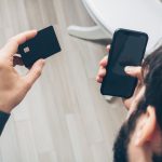 Homem segura um smartphone e um cartão de crédito enquanto realiza um pagamento online em ambiente interno.