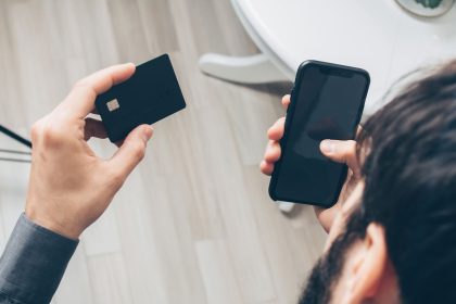 Homem segura um smartphone e um cartão de crédito enquanto realiza um pagamento online em ambiente interno.