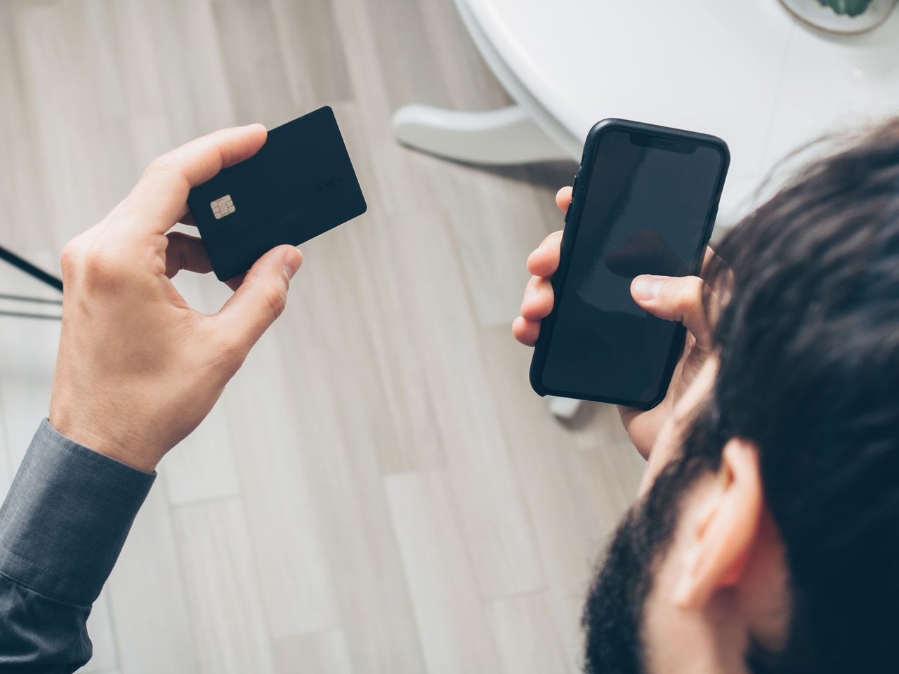 Homem segura um smartphone e um cartão de crédito enquanto realiza um pagamento online em ambiente interno.
