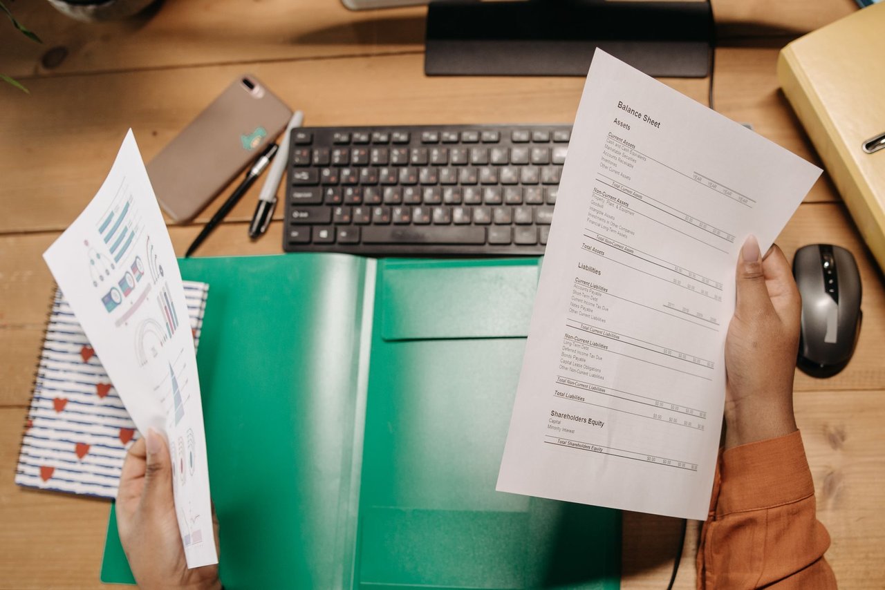 Mãos analisam planilhas financeiras sobre uma mesa com teclado e materiais de escritório.
